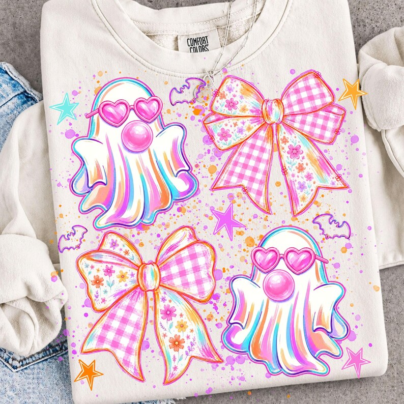May include: A cream-colored crewneck sweatshirt featuring a graphic print of two ghosts wearing pink heart-shaped glasses and two large bows with pink gingham and floral patterns, accented by small stars, bats, and colorful paint splatters.