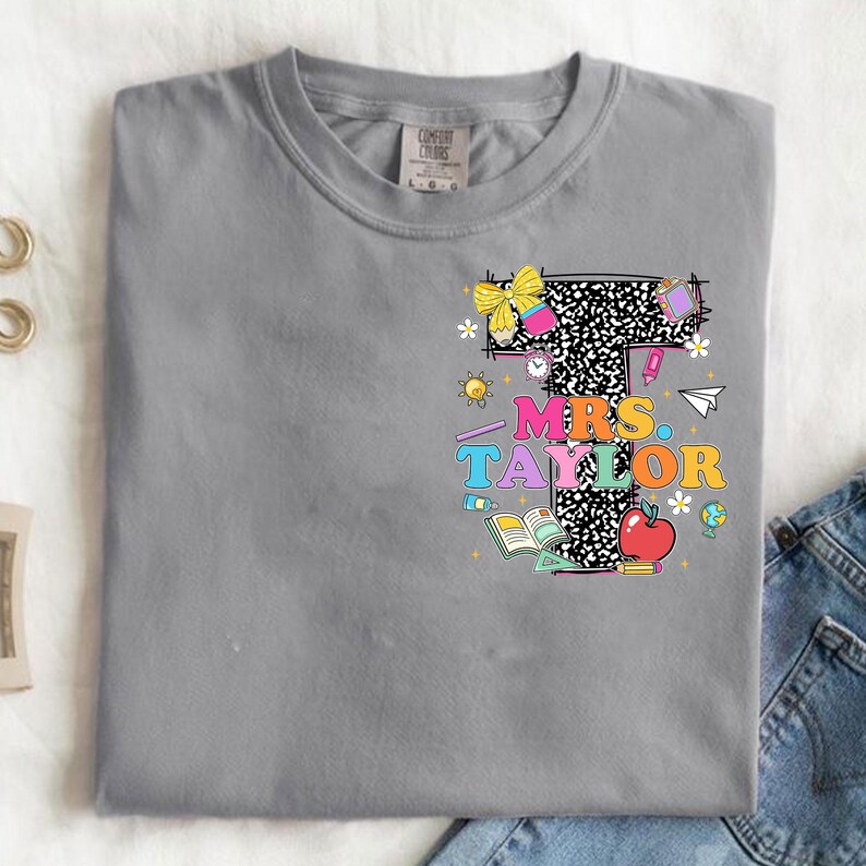 May include: A gray Comfort Colors t-shirt with a colorful graphic. The graphic features a large letter "T" with a black and white pattern, the words "MRS. TAYLOR", an apple, and school-themed illustrations.