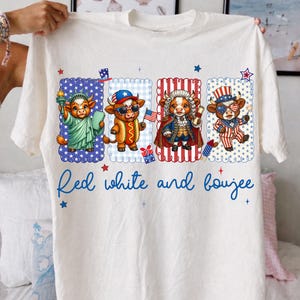 May include: White t-shirt featuring four cartoon cows dressed in patriotic attire. The cows are depicted as the Statue of Liberty, a hot dog, a historical figure, and a Fourth of July celebrant. The text "Red white and boujee" is written in blue script.