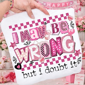 May include: White pillow with the text "I may be wrong but I doubt it" in pink and white block letters. The design includes dice, hearts, stars, and a checkered pattern. The pillow is held up by a person.