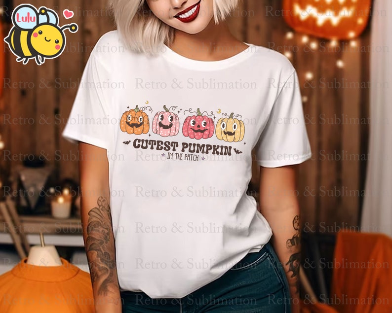 Może przedstawiać: Biała koszulka z nadrukiem trzech kresk&oacute;wkowych dyni z r&oacute;żnymi wyrażeniami twarzy. Pod dyniami znajduje się napis "Cutest Pumpkin in the Patch".