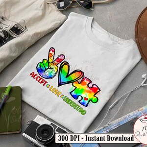 Pu&ograve; includere: Una maglietta bianca con un simbolo di pace, un cuore e un pezzo di puzzle. Il simbolo di pace e il cuore sono contornati di nero e riempiti con colori arcobaleno. Il pezzo del puzzle &egrave; riempito con un motivo tie-dye in colori arcobaleno. Il testo "ACCEPT LOVE UNDERSTAND" &egrave; scritto sotto il disegno.