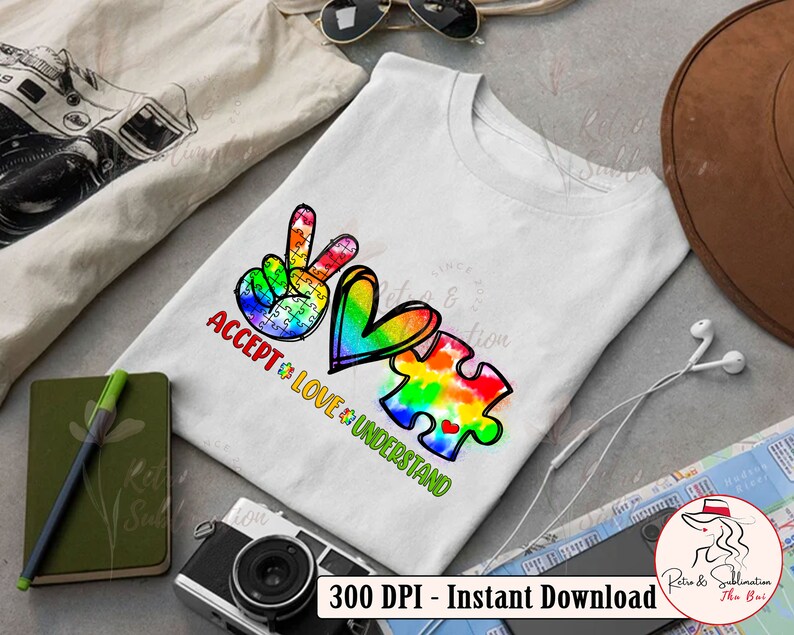Pu&ograve; includere: Una maglietta bianca con un simbolo di pace, un cuore e un pezzo di puzzle. Il simbolo di pace e il cuore sono contornati di nero e riempiti con colori arcobaleno. Il pezzo del puzzle &egrave; riempito con un motivo tie-dye in colori arcobaleno. Il testo "ACCEPT LOVE UNDERSTAND" &egrave; scritto sotto il disegno.