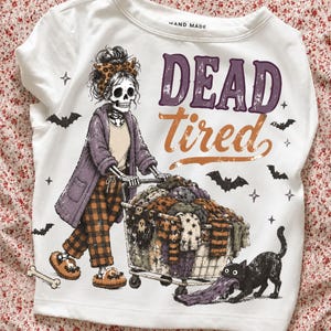 May include: A white t-shirt featuring a graphic of a skeleton wearing a leopard print headband, a purple cardigan, and plaid pants, pushing a shopping cart filled with clothes. The text reads "DEAD tired" with a black cat pulling a purple garment.