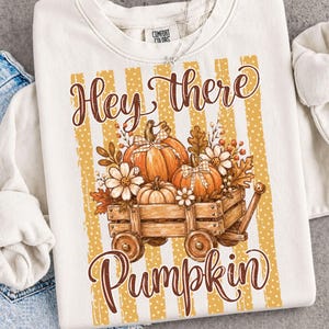 May include: A cream-colored sweatshirt featuring a graphic of a wooden wagon filled with orange pumpkins, white flowers, and autumn leaves. The text "Hey there" appears above the wagon, and "Pumpkin" is printed below in brown script.