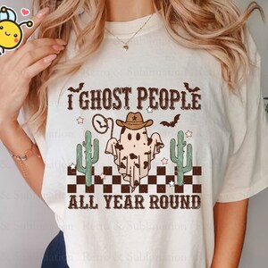 May include: A white t-shirt featuring a graphic of a ghost wearing a cowboy hat and the text "I ghost people all year round". The ghost is surrounded by cacti and stars.