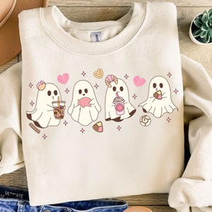 Peut inclure: Un sweat-shirt blanc avec un motif de quatre adorables fantômes tenant différents friandises. Les fantômes sont roses et blancs et ont de grands yeux. Les friandises comprennent un cochon rose, une boisson rose et blanche, une pâtisserie brune et un bonbon rose et blanc.
