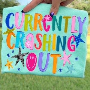 May include: A light teal t-shirt with the words "CURRENTLY CRASHING OUT" in colorful bubble letters. The shirt features various star designs, including a zebra print star and a smiley face.