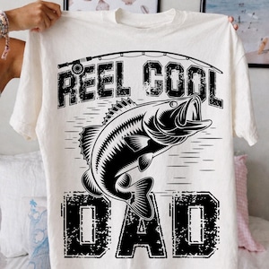 Puede incluir: Camiseta color crema con un gr&aacute;fico negro de un pez saltando fuera del agua, con las palabras "REEL COOL DAD" en una fuente desgastada. Una ca&ntilde;a de pescar est&aacute; arqueada sobre el texto. La camiseta est&aacute; sujeta por una persona.
