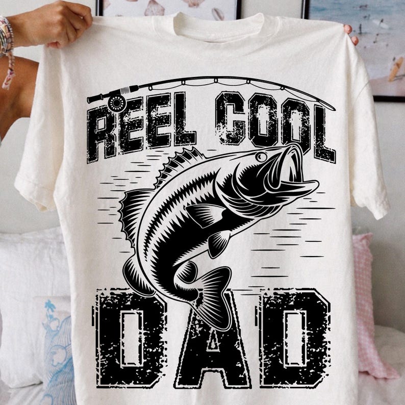 Puede incluir: Camiseta color crema con un gr&aacute;fico negro de un pez saltando fuera del agua, con las palabras "REEL COOL DAD" en una fuente desgastada. Una ca&ntilde;a de pescar est&aacute; arqueada sobre el texto. La camiseta est&aacute; sujeta por una persona.