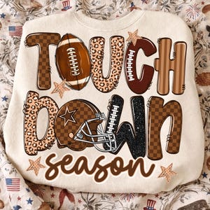 以下が含まれることがあります： 茶色、黒、ヒョウ柄の「TOUCH DOWN season」という文字がプリントされたクリーム色のスウェットシャツ。デザインには、茶色のアメリカンフットボール、茶色のアメフト用ヘルメット、文字の周りに散らばったいくつかの小さな星が含まれています。
