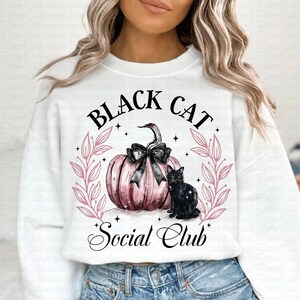 May include: White sweatshirt with a pink and black design featuring a pumpkin and a black cat. The text "Black Cat Social Club" is written in a cursive font.