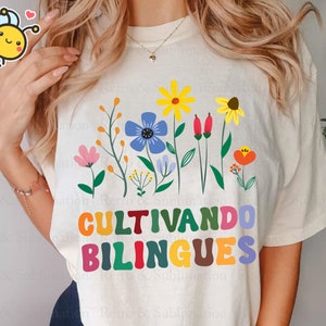Peut inclure: Un t-shirt blanc avec un motif floral color&eacute; et le texte "Cultivando bilingues" en lettres arc-en-ciel.