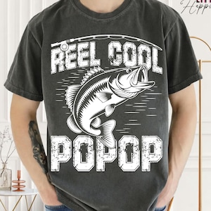 Puede incluir: Camiseta gris oscuro con un gr&aacute;fico blanco que muestra un pez saltando del agua, una ca&ntilde;a de pescar y las palabras "REEL COOL POPOP". Un estilo casual y c&oacute;modo.