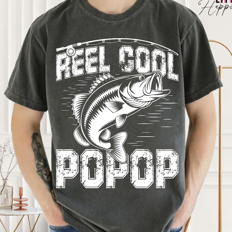 Puede incluir: Camiseta gris oscuro con un gr&aacute;fico blanco que muestra un pez saltando del agua, una ca&ntilde;a de pescar y las palabras "REEL COOL POPOP". Un estilo casual y c&oacute;modo.