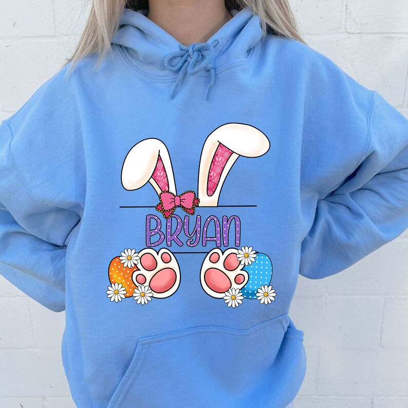 May include: Light blue hoodie with a personalized Easter bunny design. The bunny has pink ears, a pink bow, and pink paw prints. The name "BRYAN" is printed in purple on a banner.