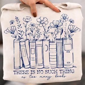 Puede incluir: Sudadera crema con un dise&ntilde;o floral y de libros azul marino. El dise&ntilde;o presenta flores y libros estilizados con el texto "THERE IS NO SUCH THING as too many books" en azul marino. La sudadera es sostenida por una mano.