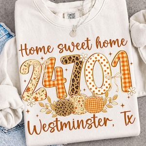 以下が含まれることがあります： Home sweet home 24701 Westminster, Txという文字がプリントされたクリーム色のクルーネックトレーナー。オレンジ色のギンガムチェック、ヒョウ柄、水玉模様で装飾された大きな数字と、3つのカボチャ、小さな花々がデザインされています。