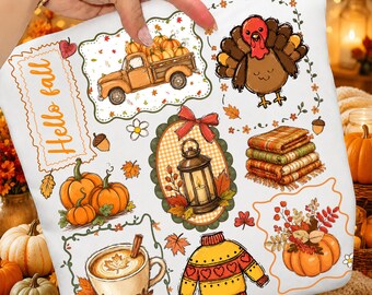 Op de afbeelding: Een wit stoffen vierkant met een kleurrijke herfstprint met een vrachtwagen vol pompoenen, een kalkoen, een lantaarn, dekens, een koffiemok en een gele trui. De tekst "Hello fall" is in oranje letters op de stof gedrukt.