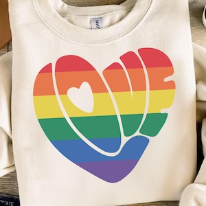 Pode incluir: Uma camisa de moletom branca com um design de coração arco-íris que soletra "LOVE" em uma fonte retrô.