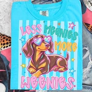 Könnte beinhalten: Hellblaues T-Shirt mit einem Cartoon-Dackel, der eine herzförmige Sonnenbrille trägt. Der Text lautet "LESS MEANIES MORE WEENIES". Der Hund ist braun und hellbraun. Das T-Shirt hat blaue und weiße Streifen.