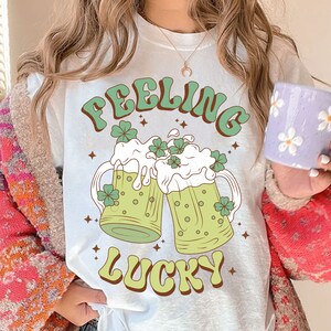 May include: White t-shirt with a green and white graphic of two beer mugs with shamrocks and the text "Feeling Lucky".