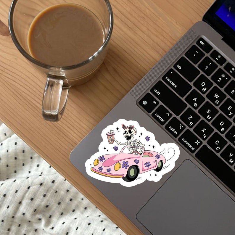May include: A sticker featuring a cartoon skeleton wearing sunglasses and driving a pink car with purple flowers. The skeleton is holding a pink and white cup with a straw.