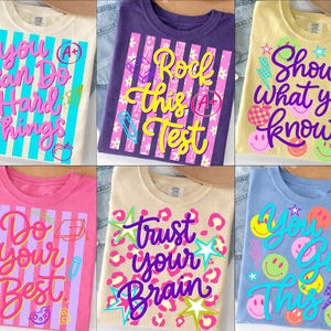 May include: Six colourful t-shirts with school-themed graphics. Each shirt features a different motivational phrase in a playful font, such as "You Can Do Hard Things" and "Show What You Know", along with related illustrations.