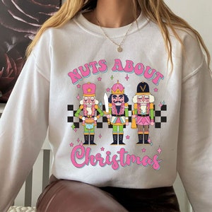 May include: White sweatshirt with a pink and blue graphic of three nutcracker figurines playing instruments. The text "NUTS ABOUT CHRISTMAS" is written in pink letters above the figurines.
