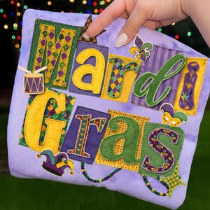 May include: A lavender fabric item with the words "Mardi Gras" appliquéd in various colours and patterns. The letters are yellow, green, purple, and gold, with decorative elements like a drum, fleur-de-lis, and a jester's hat.