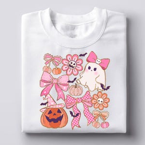 May include: White sweatshirt featuring a colorful Halloween-themed design. The design includes a smiling jack-o'-lantern, pink bows, flowers, a ghost, pumpkins, bats, and cherries. The graphics are in shades of pink, orange, and gold.