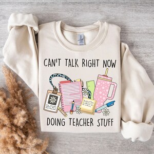 Puede incluir: Una sudadera beige con el texto "Can't talk right now doing teacher stuff" impreso en ella. El texto está en negro y hay un gráfico de útiles escolares en rosa, verde y azul.