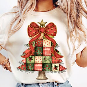 May include: A white t-shirt featuring a red and green patchwork Christmas tree design. The tree has a large red bow with gold accents and a gold star on top.