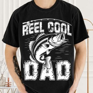 Puede incluir: Camiseta negra con un gr&aacute;fico blanco de un pez y una ca&ntilde;a de pescar. El texto de la camiseta dice "REEL COOL DAD" en una fuente desgastada. La camiseta es de cuello redondo y tiene mangas cortas.