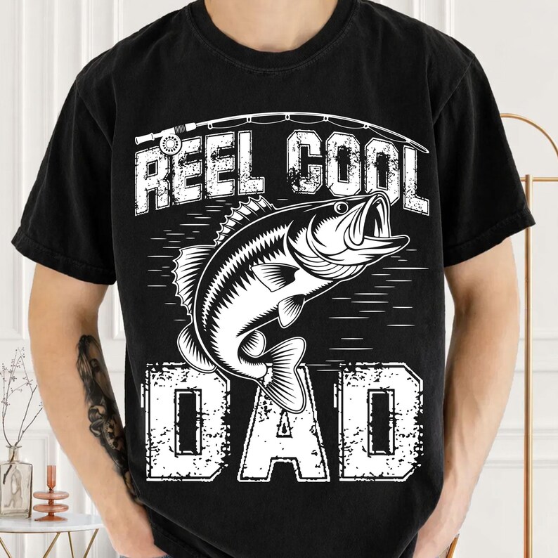 Puede incluir: Camiseta negra con un gr&aacute;fico blanco de un pez y una ca&ntilde;a de pescar. El texto de la camiseta dice "REEL COOL DAD" en una fuente desgastada. La camiseta es de cuello redondo y tiene mangas cortas.