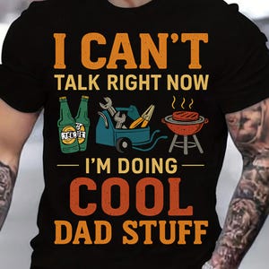 May include: Black t-shirt with a vintage-style graphic that says "I can't talk right now I'm doing cool dad stuff". The graphic features a toolbox, a grill, and two bottles of beer.