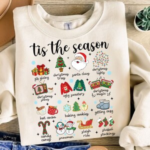 May include: A white sweatshirt with the text "tis the season" in red and black lettering. The sweatshirt features a variety of Christmas-themed illustrations, including a Christmas tree, Santa Claus, a stocking, a snowman, and a sleigh.