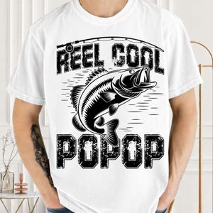 Puede incluir: Camiseta blanca con un gr&aacute;fico negro de un pez saltando del agua, una ca&ntilde;a de pescar y las palabras "REEL COOL POP POP". El dise&ntilde;o est&aacute; centrado en la camiseta.