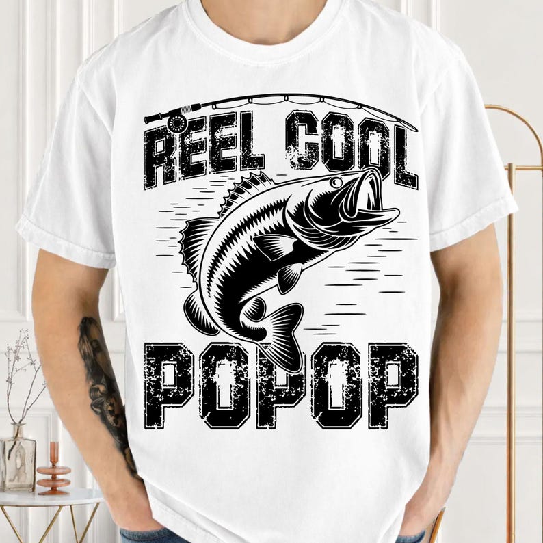 Puede incluir: Camiseta blanca con un gr&aacute;fico negro de un pez saltando del agua, una ca&ntilde;a de pescar y las palabras "REEL COOL POP POP". El dise&ntilde;o est&aacute; centrado en la camiseta.