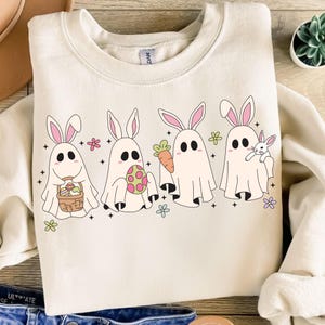 Puede incluir: Una sudadera blanca con un gráfico de cuatro conejos fantasma sosteniendo artículos con temática de Pascua. Los conejos llevan orejas de conejo rosas y tienen cuerpos de fantasmas blancos. Están sosteniendo una cesta de huevos, una zanahoria, un huevo de Pascua y un conejito pequeño.