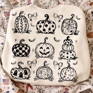 May include: A cream-colored crewneck sweatshirt featuring a black ink-style graphic print of nine distinct pumpkins. The designs include patterns like polka dots, spiderwebs, stars, flowers, ghosts, and a jack-o'-lantern, accented by small bows and bats.