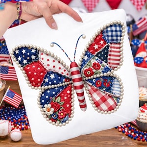 Op de afbeelding: Wit vierkant kussen met een patriottisch vlinderontwerp. De vleugels van de vlinder zijn patchwork met rode, witte en blauwe stoffen, versierd met parel kralen. Het lichaam van de vlinder is rood en wit gestreept. Het kussen wordt door een persoon vastgehouden.