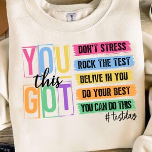 Op de afbeelding: Een wit sweatshirt met een kleurrijke afbeelding die "You got this" zegt en de tekst "Don't stress, Rock the test, Believe in you, Do your best, You can do this, #testday" bevat.