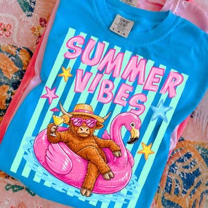 May include: A turquoise t-shirt with the words "SUMMER VIBES" in pink. The graphic shows a Highland cow wearing sunglasses and a straw hat, relaxing on a pink flamingo inflatable, holding a cocktail. The t-shirt is folded.