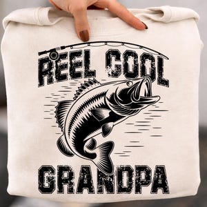 Puede incluir: Sudadera color crema con un gr&aacute;fico negro. El gr&aacute;fico presenta un pez saltando fuera del agua, una ca&ntilde;a de pescar y las palabras "REEL COOL GRANDPA" en una fuente desgastada. La sudadera es sostenida por una persona.