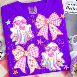 May include: A purple t-shirt featuring a graphic print of two white ghosts wearing pink heart-shaped sunglasses and blowing bubblegum, accompanied by two large pink gingham and floral bows, small pink and blue stars, and bats.