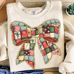 May include: A cream-colored sweatshirt with a large, colorful patchwork bow printed on the front. The bow is outlined in gold glitter and has a red, green, blue, and white color scheme.
