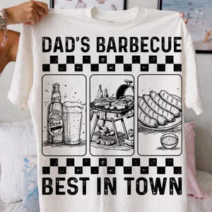 May include: Off-white t-shirt with a black and white graphic. The design includes the text "DAD'S BARBECUE BEST IN TOWN" and three illustrated panels: a beer bottle and glass, a barbecue grill with food, and a plate of sausages.