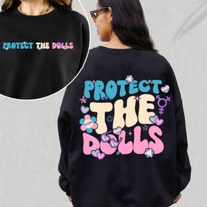 Może przedstawiać: Czarna bluza z napisem "Protect the Dolls" w kolorach tęczy i kwiatem z napisem "Pride".