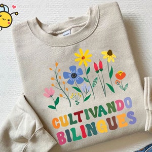 Peut inclure: Un sweat-shirt beige avec un motif floral color&eacute; et le texte "Cultivando bilingues" en lettres arc-en-ciel.